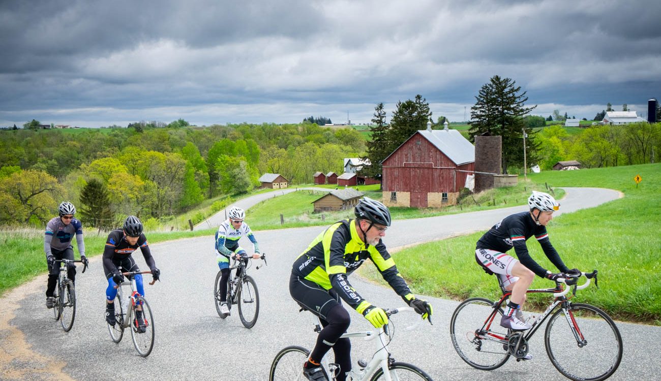 Five riders pedal up a hill on a curvy road with a red barn behind them.