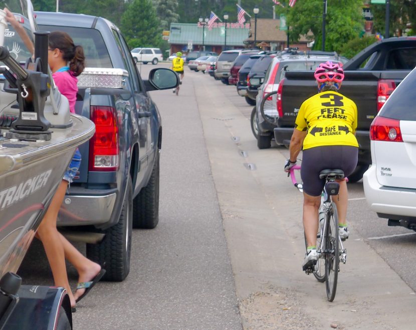 Cyclists wearing 3ft jerseys ride between parked cars and a pickup pulling a boat on a trailer