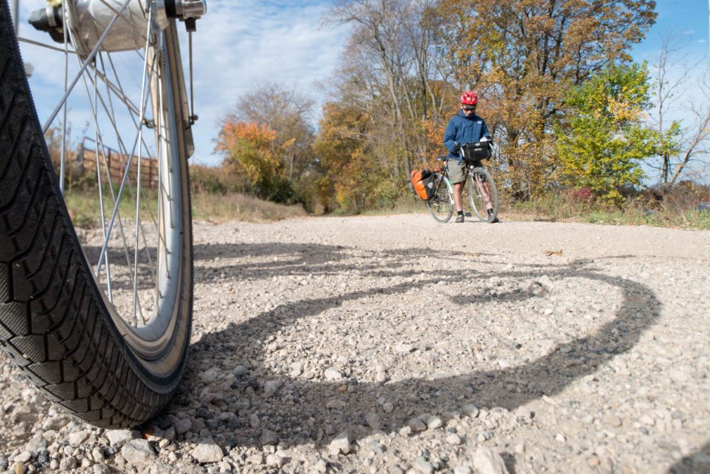 Bicycle tire in foreground with person on bike in background on gravel trail