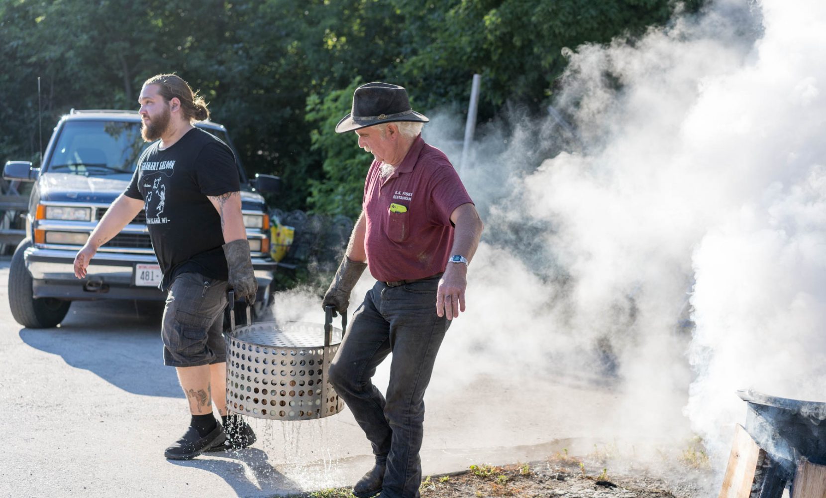 Two men carry fish boil pot away from fire.