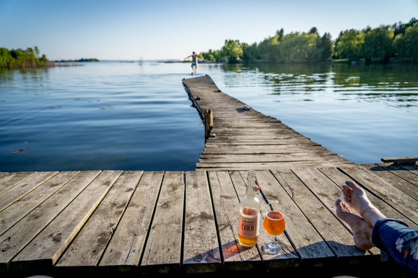 Bottle and glass of cider next to crossed legs and bare feet on wooden pier on Washington Island with boy jumping into water in distance.