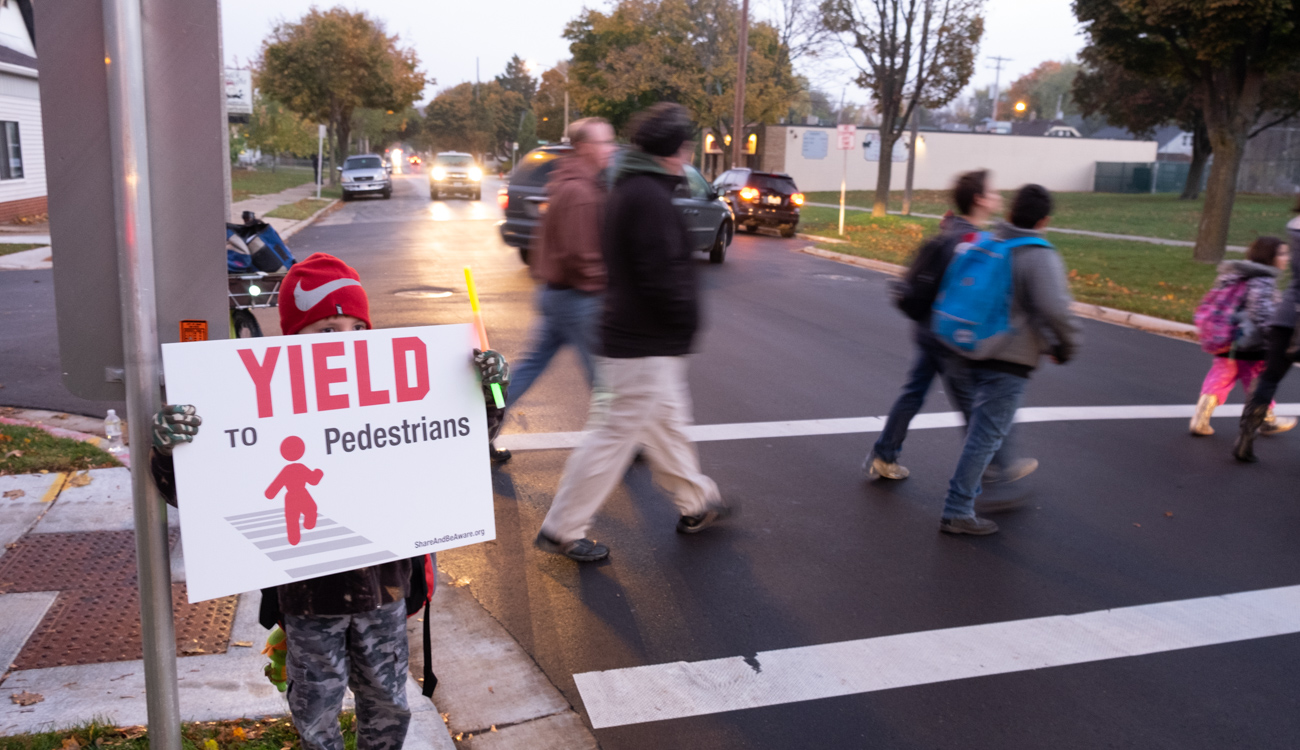 child holds Yield to Pedestrians sign in front of people in crosswalk behind him.