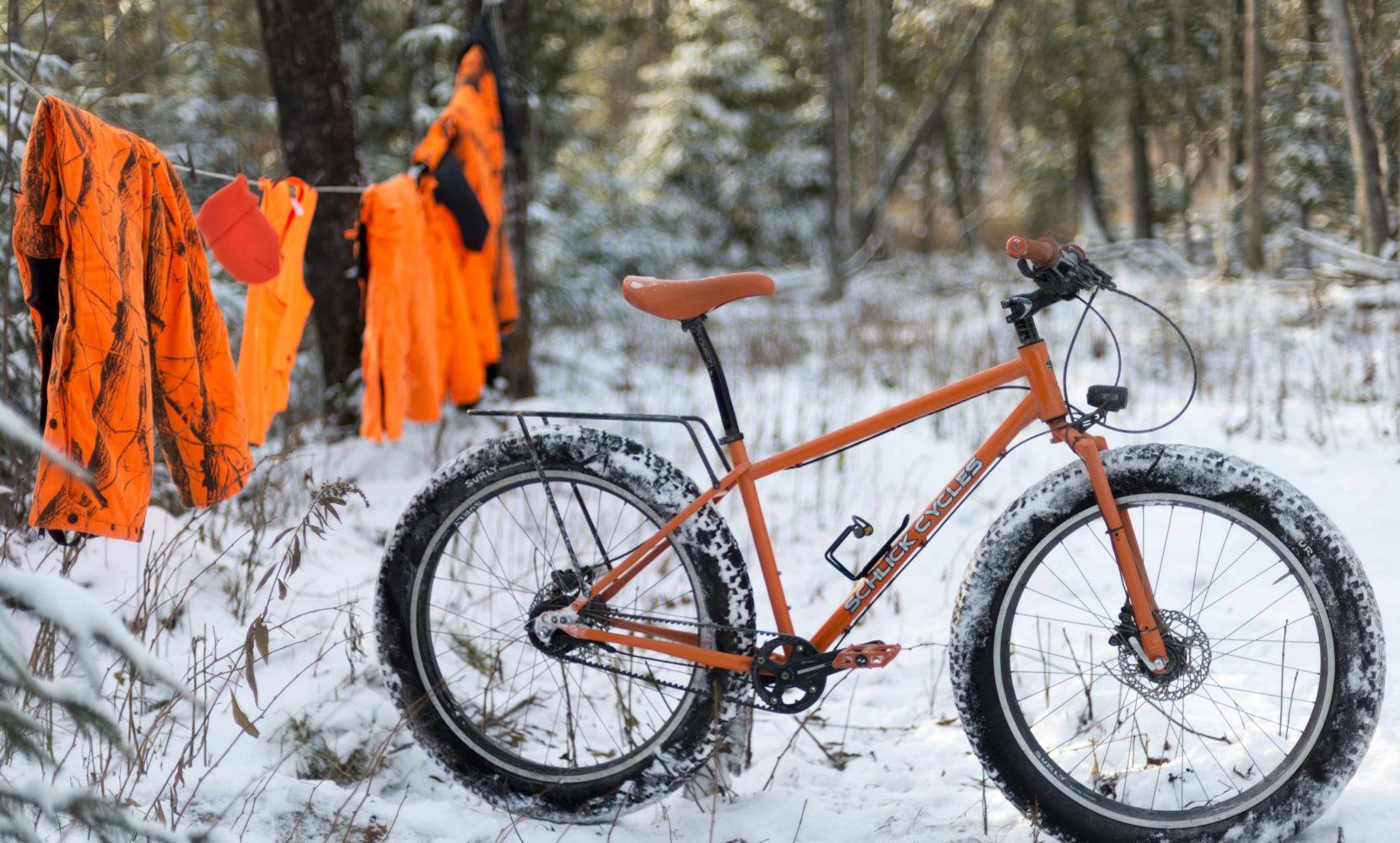 Orange Schlick Northpaw fatbike in snow by deer unting cltohs hangin gon line in woods