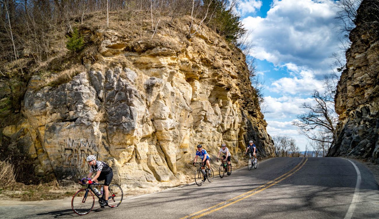 Four cyclists riding through a hand hewn cut in a limestone bluff