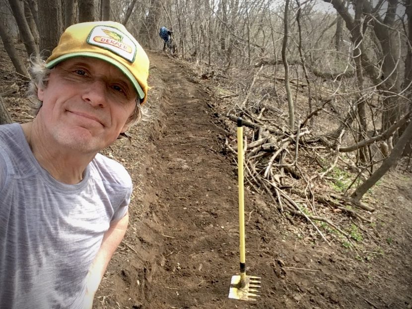 Man in Dekalb hat poses in front of a Mcloed tool on a bench cut trail with a bike in background