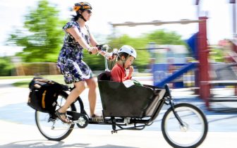 Woman riding a cargo bike with child in front past a playground