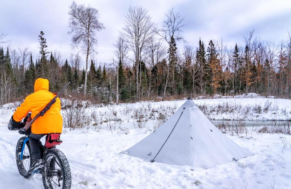 Man on bicycle with rifle over his shoulder rides in snow past tent.
