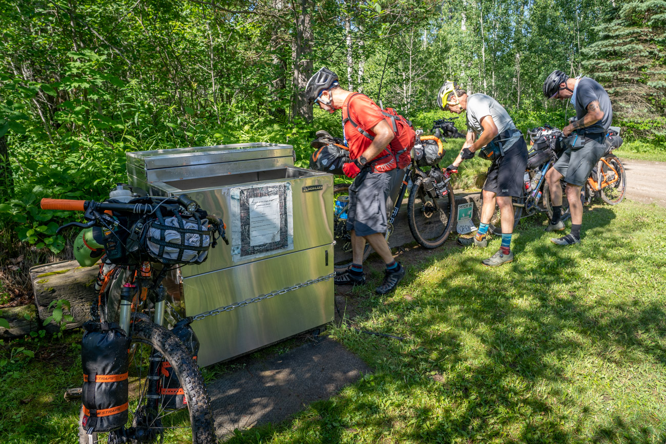 An open refridgerated cooler in the forest next to three people with bicycles