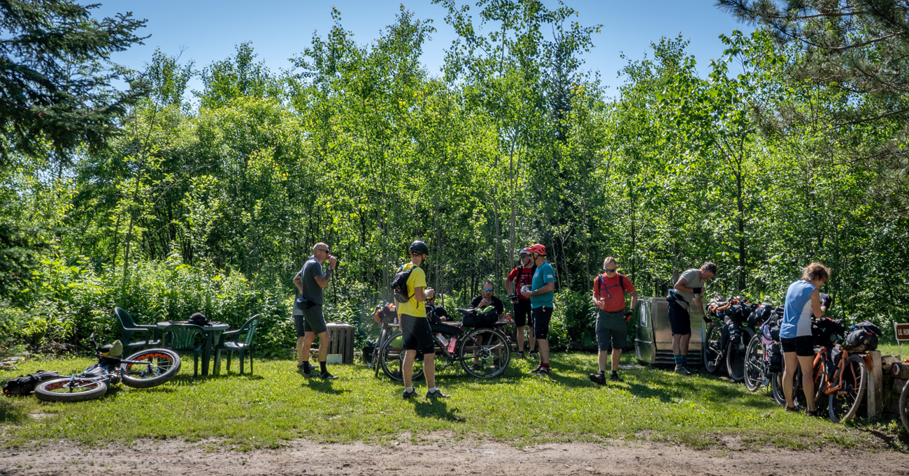 A group of cyclists next to tables, chairs and a cooler on the side of a gravel road