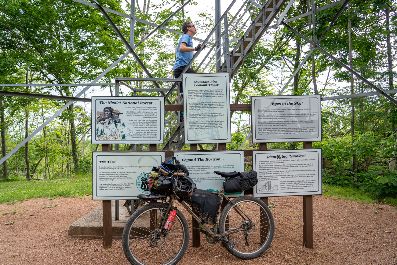 Man climbs the stairs at base of firetower with informational signs and bicycle below