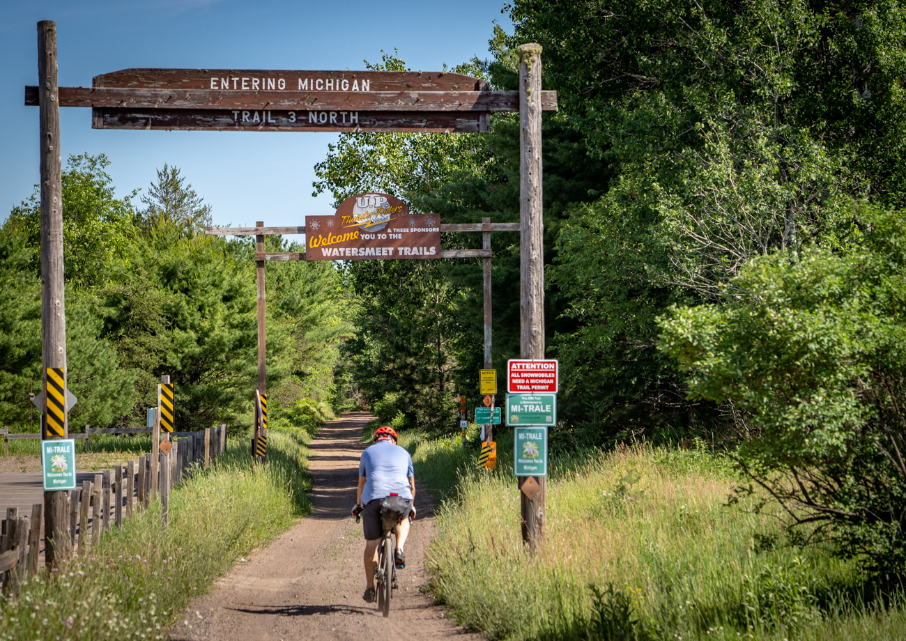Man rides bicycle onto trail under entering Michigan sign