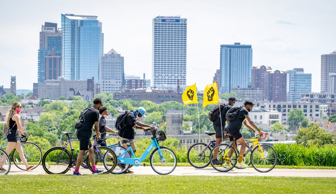 People walk bikes behind two black power flags in front of Milwaukee skyline