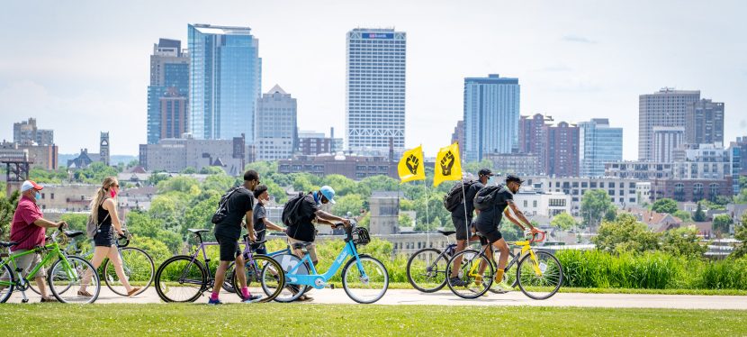 People walk bikes behind two black power flags in front of Milwaukee skyline