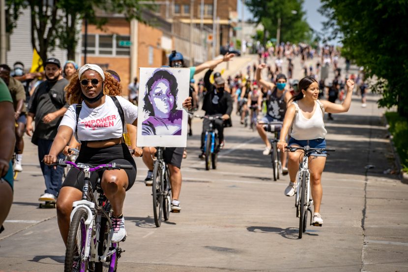 A woman riding a bike in the Black is Beautiful ride holds up a poster of Breonna Taylor