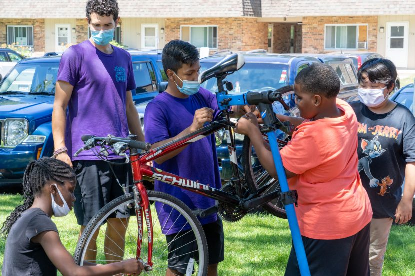 A group of kids work on a bicycle in a repair stand while two instructors help. The instructors have maskes, some kids do others do not.