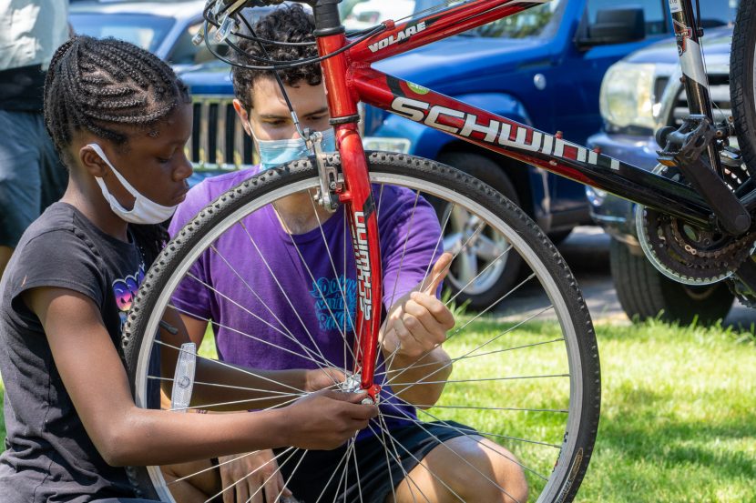 A man wearing a mask helps a girl install a front bicycle wheel.