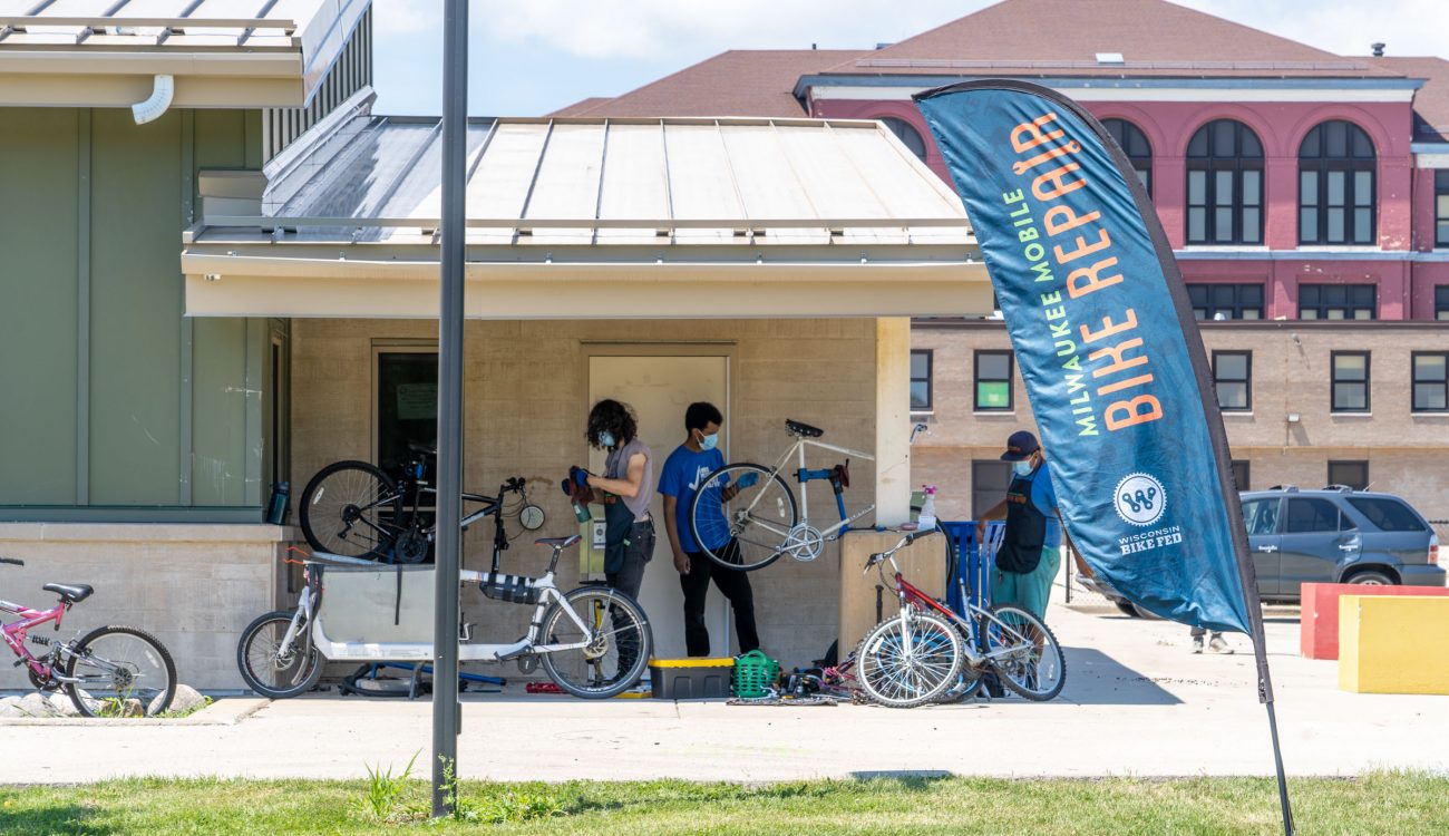 Three people wearing masks work on bicycles under an awning with a Mobile Bike Repair flag in the foreground.