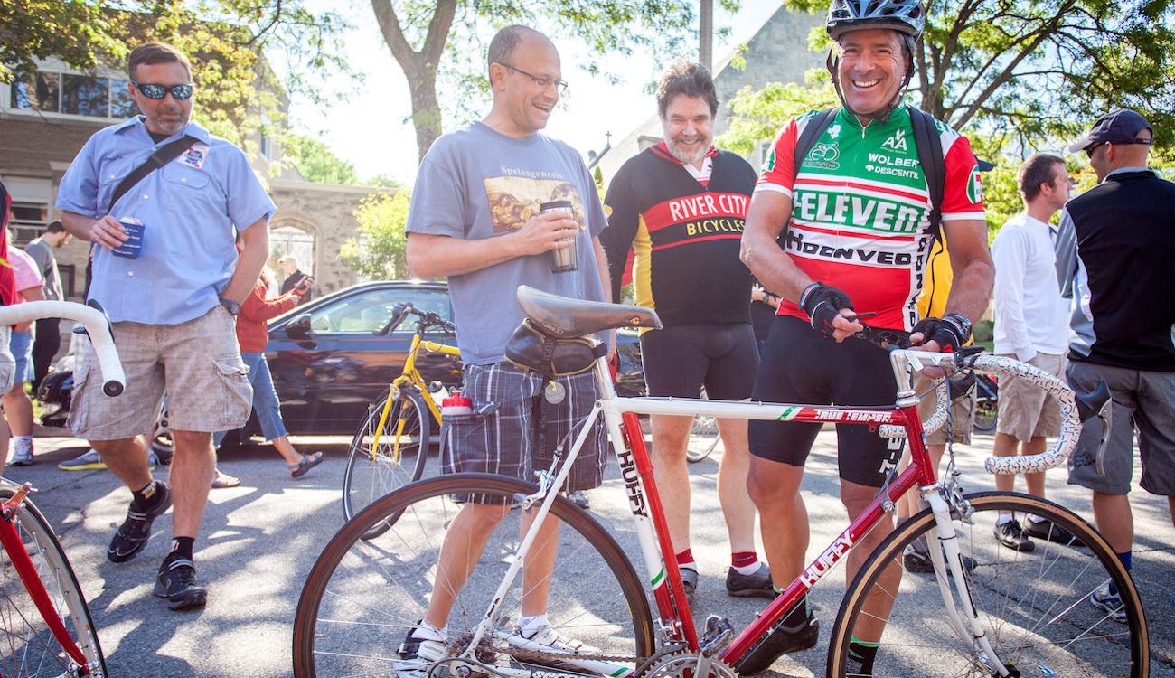 Tom Schuler in 7-Eleven jersey stands next to his 7-Eleven Team Bike with others, laughing.