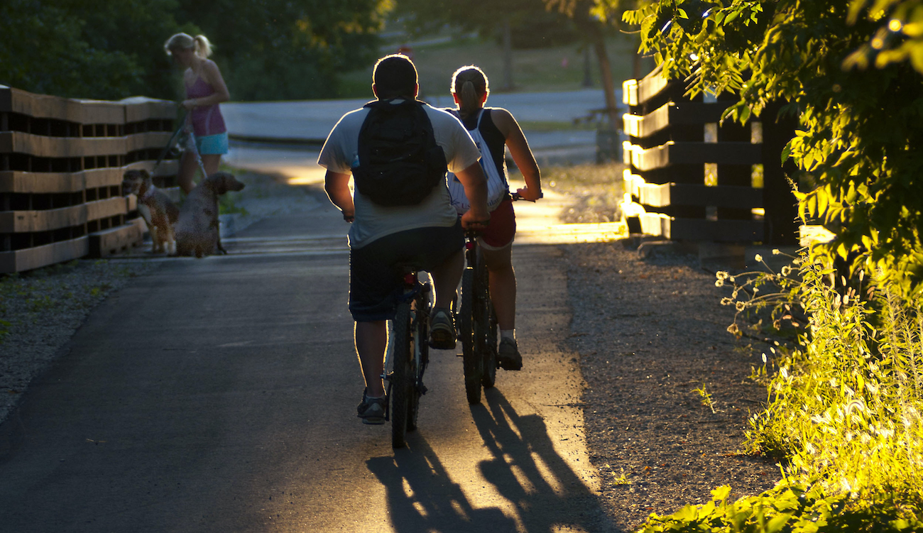 backlit photo of two kids riding bikes on trail and woman with dog on leash