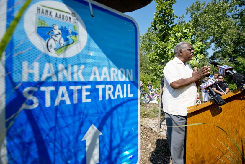 Hank Aaron speaks at a podium with news microphones and a Hank Aaron State Trail sign in the foreground.