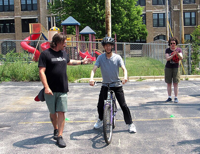 man wearing grey shirt and helmet practices learning to ride ona n adult sized balance bike without pedals in a parking lot with safety instructor looking on