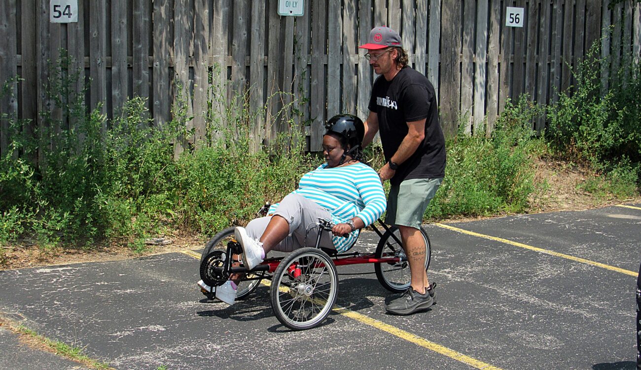 woman pedals an recumbent tricycle in parking lot with help of adult male safety instructor