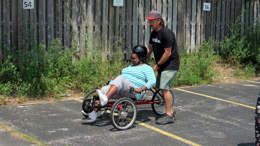woman pedals an recumbent tricycle in parking lot with help of adult male safety instructor