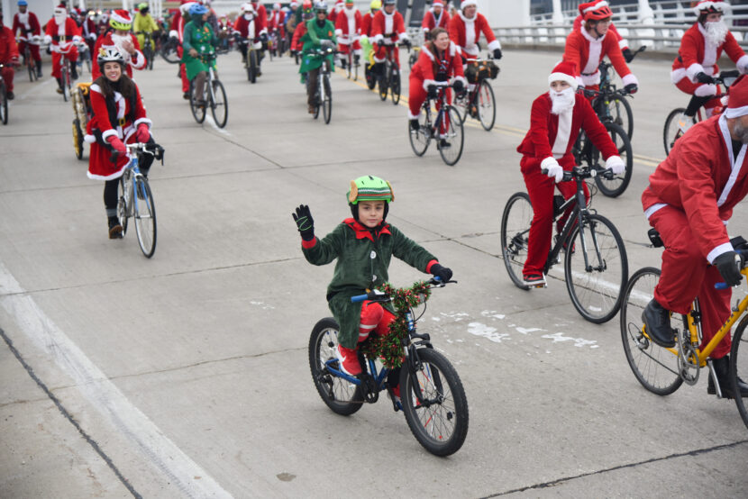 large group of costumed riders pedal the streets. In front is a young kid dressed as an elf and waving