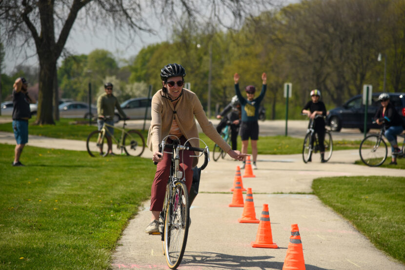 A person on a bike navigates a course marked with cones while an instructor signals behind them.