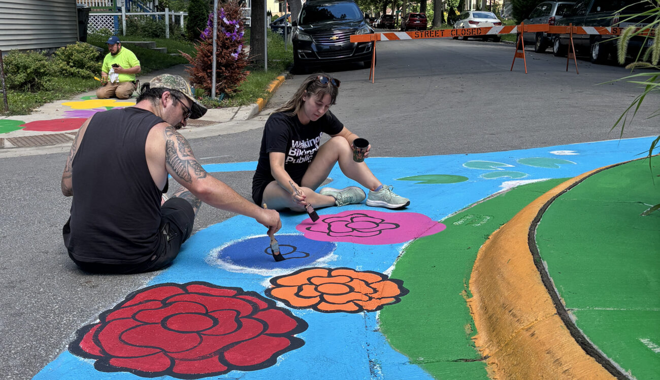 Two people sit on the street painting a colorful design near a traffic circle.