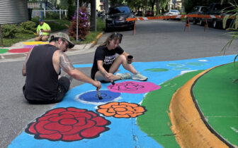 Two people sit on the street painting a colorful design near a traffic circle.