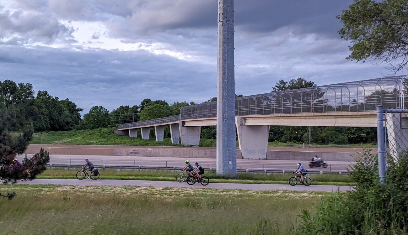 A group of bicyclists bike alongside the Beltline in Madison with the Cannonball path bridge in the background.