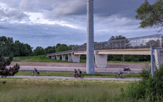 A group of bicyclists bike alongside the Beltline in Madison with the Cannonball path bridge in the background.